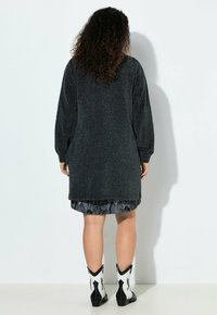Maglione grigio oversize con maniche lunghe, abbinato a una gonna fantasia e stivali da cowboy in bianco e nero. Sfondo bianco semplice.