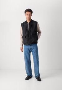 Mann steht an einer weißen Wand, trägt eine schwarze Weste, ein beiges Langarmhemd, blaue Jeans und schwarze Slipper mit einer Hand in der Tasche.