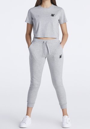 Mujer que lleva una camiseta corta gris claro y pantalones jogger a juego con detalles de logo en negro y zapatillas blancas, con las manos en los bolsillos.