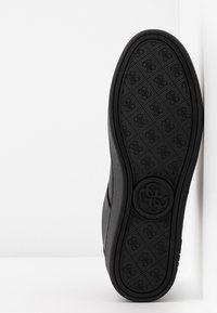 Suola di scarpa nera con battistrada a motivo e emblema circolare, posizionata verticalmente contro un angolo di muro bianco.