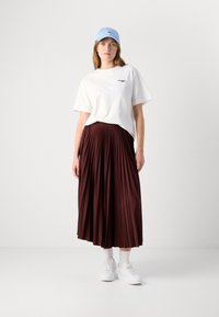 Hvit bomull T-skjorte med grønn logo, kombinert med en burgunder plissémidi-skjørt. Tilbehøret inkluderer en lyseblå caps og hvite sneakers.