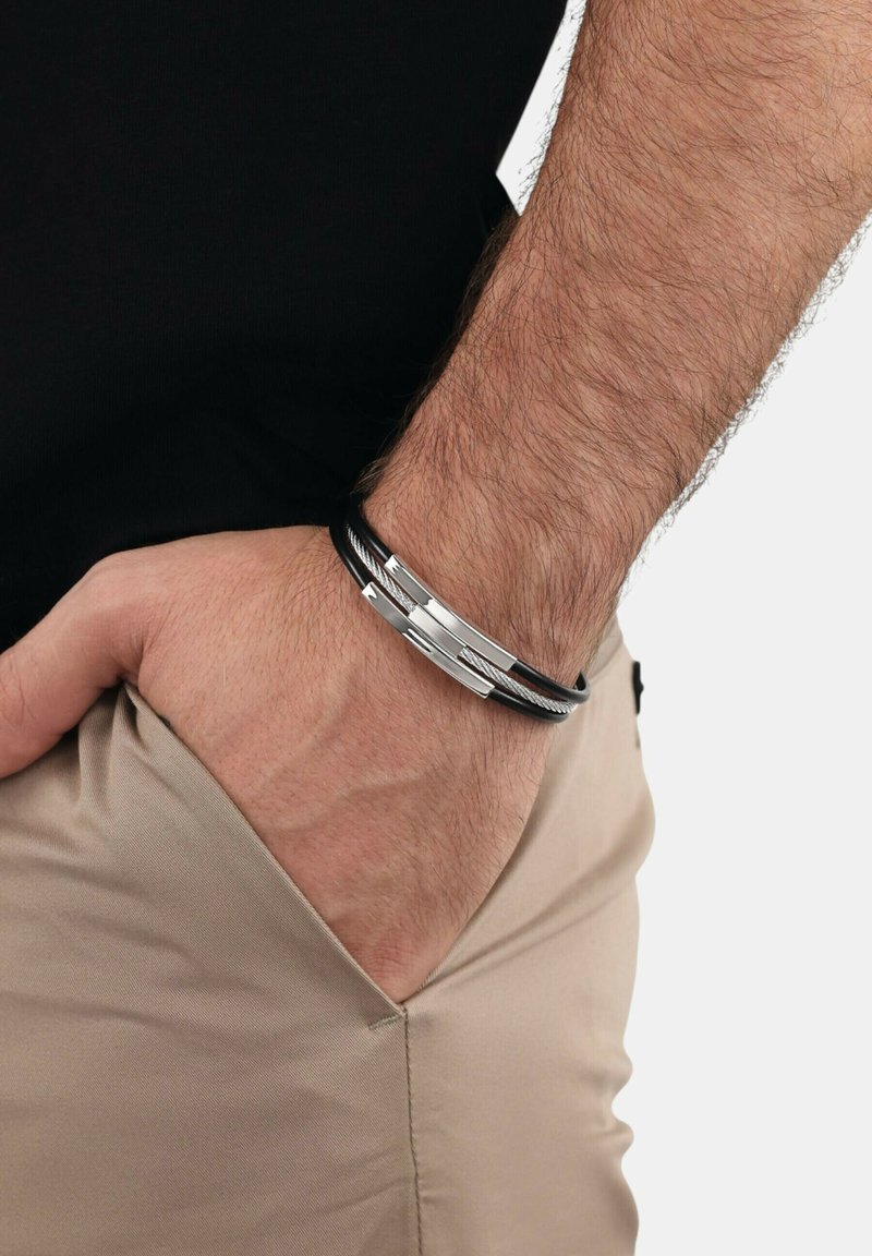 Bracciale in pelle nera con accenti metallici argentati. Presenta un design stratificato e dettagli della chiusura. Indossato al polso, abbinato a pantaloni beige.