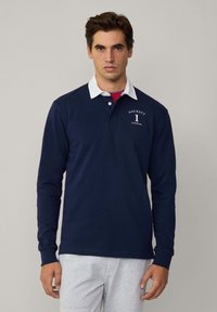 Mornarska polo majica dugih rukava s bijelim ovratnikom, sa logotipom "Hackett 1 London" na lijevoj strani grudi i rebrastim manžetama.