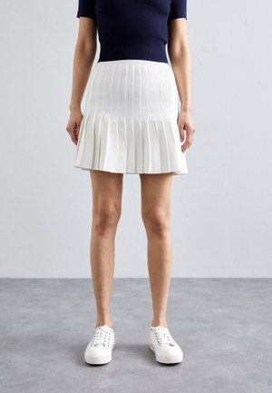 Vrouw in een witte plooienrok, marineblauw T-shirt met korte mouwen en witte sneakers, staande tegen een effen lichtgrijze achtergrond.