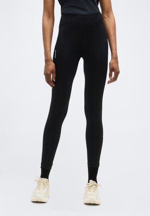 Personne portant des leggings noirs taille haute avec un logo blanc sur la cuisse, debout les bras détendus, baskets beiges à lacets, fond clair uni.