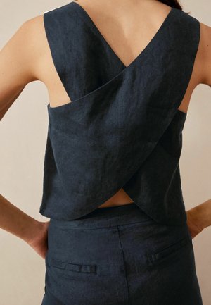 Vrouw met een mouwloze marineblauwe top met brede gekruiste bandjes en bijpassende broek met hoge taille, handen op de heupen, achteraanzicht.