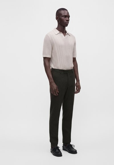 Man wearing a beige short-sleeve knit polo shirt, dark green tailored pants, and black lace-up shoes standing against a plain background.