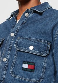 Giacca di jeans di colore blu scuro con bottoni argentati, due tasche sul petto e un'etichetta del logo Tommy Jeans con colori rosso, bianco e verde.