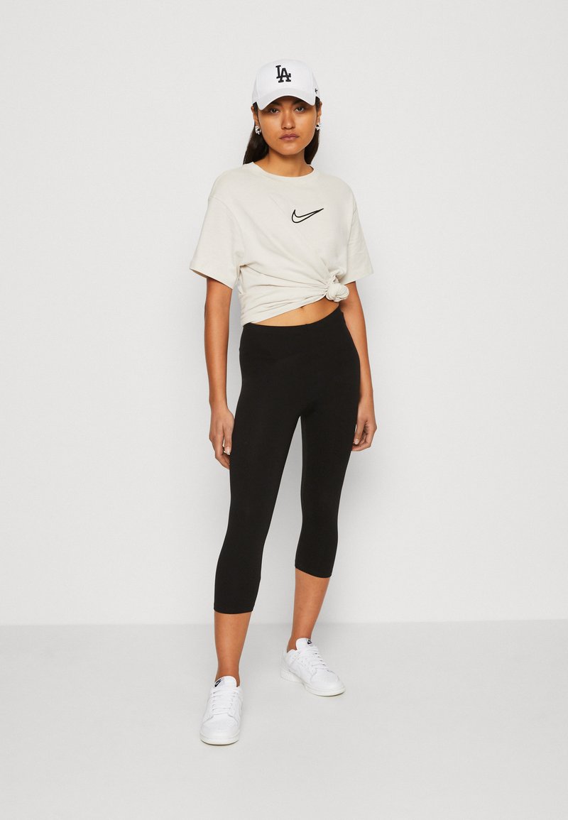 Fekete cropped leggings egy csomóra kötött bézs pólóval, amelyen fekete Nike logó található. Egy fehér sapka teszi teljessé az sportos megjelenést.