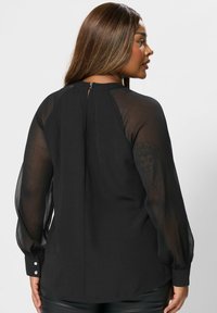 Blusa nera con scollo alto, maniche lunghe trasparenti e polsini con bottoni; presenta una chiusura a chiave sul retro e una texture liscia.