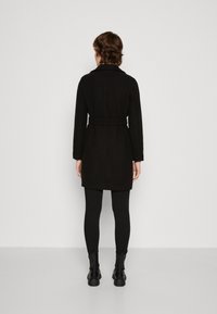 Svart skräddarsydd kappa med krage, bälte i midjan och långa ärmar. Bärs över svarta leggings och ankelboots, slät textur, minimalistisk design.