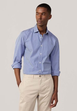 Camisa de botones a rayas verticales azul y blanca; mangas largas con puños enrollados. Combinada con pantalones beiges, mostrando un corte estándar.