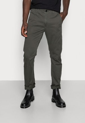 Mężczyzna w ciemnozielonych podwiniętych spodniach i czarnych skórzanych botkach, stojący z rękami w kieszeniach na jednolitym tle.