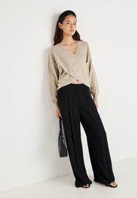 Pulover bej tricotat cu guler în V și model în carouri diagonale, asociat cu pantaloni largi negri și o geantă de mână neagră.