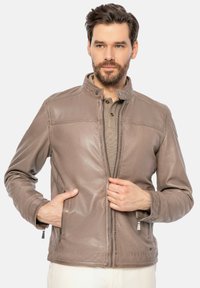 Giacca in pelle marrone con colletto alto, tasche con zip, e una texture liscia. Presenta dettagli cuciti e un design verticale.