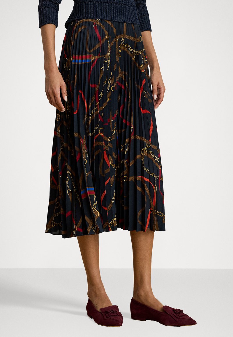 Fustă plisată de culoare închisă, cu un model de lanț și panglici în roșu și auriu, asociată cu mocasini din suede burgundy cu un accent de fundiță.