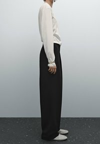 Person som står på sidan iförd en vit långärmad tröja, svarta vida byxor, vita skor och guldarmband på handleden.