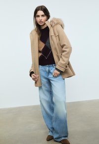 Cappotto in misto lana beige con cappuccio foderato in pelliccia, abbinato a un maglione a blocchi di colore scuri e a jeans a gamba larga di un blu chiaro. Stivaletti alla caviglia marroni.