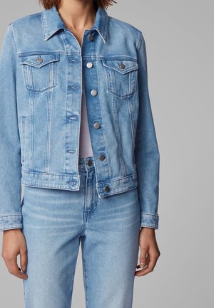 Person trägt eine hellblaue Jeansjacke mit Knöpfen und passende Jeans vor einem einfarbigen grauen Hintergrund.