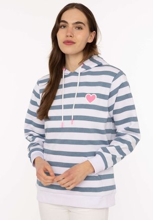 Junge Frau trägt einen weiß-grau gestreiften Hoodie mit einem pinken Herz auf der Brust und steht vor einem schlichten weißen Hintergrund.