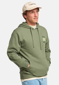 Muž nosiaci béžovú čiapku a olivovo zelenú mikinu s predným vreckom, v kombinácii so svetlomodrými džínsami, ruky v vreckách, pozerá sa doprava.