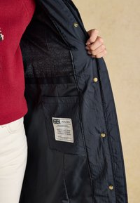 Person wearing red sweater holds open navy jacket showing inner pocket with a Joules brand label and gold button details.
