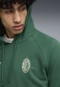 Groene hoodie met een trekkoord, voorzien van een crèmekleurig embleem met de tekst "ACM 1899" op de borst. Gemaakt van een zachte, gestructureerde stof.