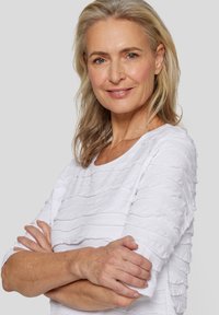 Weißes Langarmoberteil mit strukturierten Rüschen, runder Ausschnitt und lässiger Passform, getragen von einer Frau mit hellbraunem Haar und natürlichem Makeup.