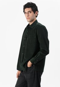 Dark green corduroy shirt featuring a button-up design, chest pocket, and subtle ribbed texture. Worn with dark jeans.