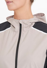 Lätt beige vindjacka med svarta detaljer. Har en figursydd huva, framzipper och reflekterande vit rand på axlarna.