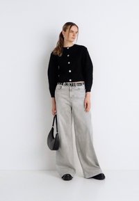 Μαύρο πλεκτό ζακέτα με στρογγυλά κουμπιά, συνδυασμένο με φαρδιά γκρι ανοιχτή τζιν και μαύρα παπούτσια, κρατώντας μια μικρή μαύρη τσάντα.