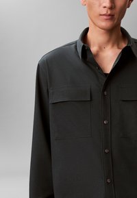 Camicia verde scuro con bottoni, due tasche sul petto, colletto classico e tessuto liscio e strutturato. Presenta dettagli di cuciture sobrie.