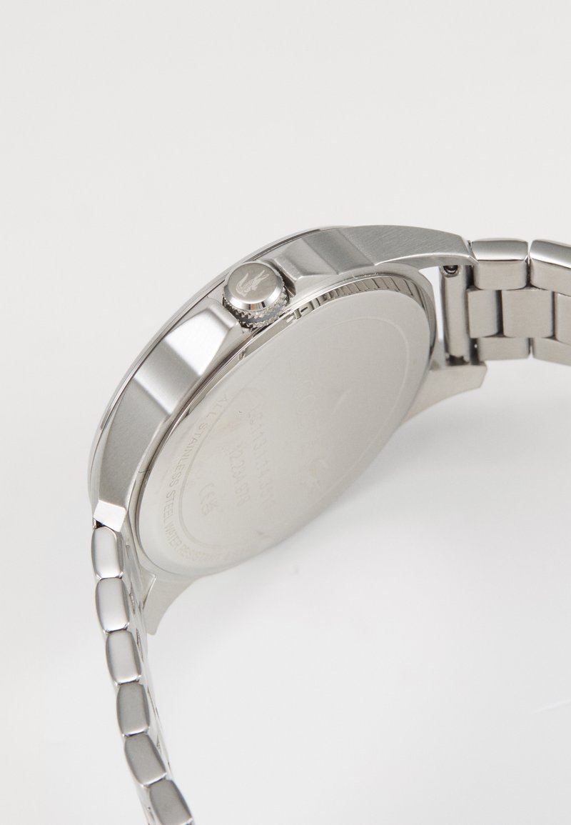 Reloj de acero inoxidable plateado con una caja redonda pulida, corona texturizada y una pulsera de eslabones. La parte trasera está grabada con especificaciones.