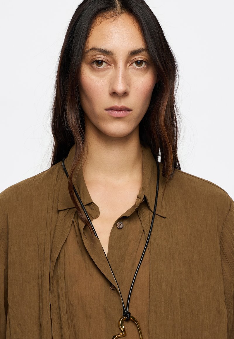 Camisa marrón texturizada con un detalle de botón y un collar de cordón negro con un colgante abstracto dorado. Elementos de diseño simples y naturales.