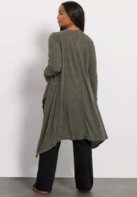 Długi kardigan z otwartym przodem w oliwkowym kolorze z teksturowanym wzorem dzianiny. Ma długie rękawy i swobodną sylwetkę, noszony na luźne czarne spodnie.