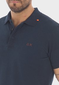Polo shirt blu navy con colletto, caratterizzato da accenti ricamati arancioni e un sottile logo geometrico sul petto. Tessuto morbido e testurizzato.