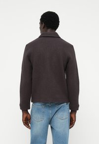 Brown wool-blend jacket with a collar, long sleeves, and a straight cut. Worn over light blue denim jeans, features a textured surface.