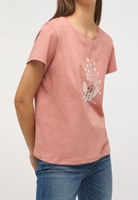 Camiseta de algodón rosa con mangas cortas y cuello redondo. Presenta una impresión gráfica de flores y pájaros en el frontal en blanco y azul.