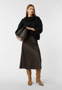 Black knitted sweater with a cowl neck, brown satin midi skirt, and tan suede knee-high boots. Model holds a black woven bag.