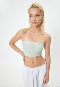 Jasnozielony, prążkowany top typu bandeau z detalami teksturowymi; krótki fason. Modelka nosi go z białymi spodniami na elastycznej talii.