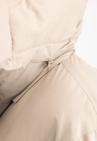 Veste beige avec capuche fixée par une fermeture éclair, gros plan sur la texture du tissu et les coutures au niveau de l'épaule.