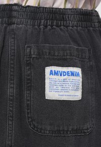 Fekete denim rövidnadrág rugalmas derékkal, nagy fehér címkével, kék szöveggel a hátsó zsebében. Texturált anyag, laza dizájn.