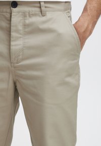 Beige cotton trousers with a smooth texture, featuring a button closure and side pockets. Tailored fit with visible seams and minimal detailing.