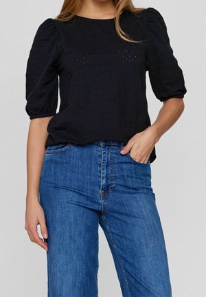 Femme portant un chemisier noir à manches bouffantes texturées et un jean taille haute bleu, avec la main droite dans la poche avant.