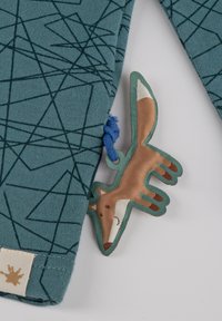 Türkiser Pullover mit abstraktem, dunkelgrünem geometrischen Muster. Verfügt über ein braunes Stoff-Fuchs-Schild mit einer blauen Schnur.