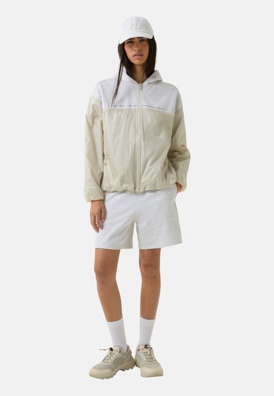 Beige windbreaker met grijze bovenpaneel, ritsluiting, en een koord aan de onderkant; gecombineerd met grijze shorts en tan sneakers. Een witte pet maakt de look compleet.