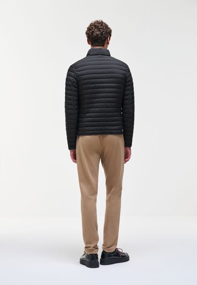 Homme portant une veste matelassée noire, un pantalon beige et des chaussures noires, debout dos à la caméra sur un fond blanc.