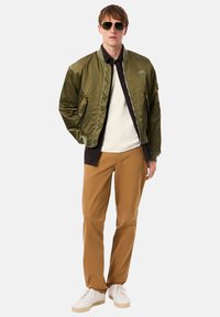 Veste bomber vert olive à finition brillante, associée à une chemise blanche et un pantalon marron. Des lunettes de soleil et des baskets beige complètent le look.