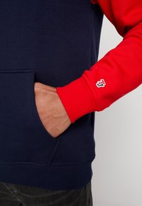 Sudadera con capucha que presenta un cuerpo azul marino y mangas rojas, un bolsillo canguro, puños acanalados y un pequeño logo bordado en la manga.