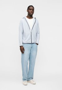 Άνδρας που στέκεται φορώντας γκρι ανοιχτό φούτερ με φερμουάρ, λευκό μπλουζάκι, ανοιχτό μπλε τζιν και λευκά αθλητικά παπούτσια μπροστά σε απλό φόντο.